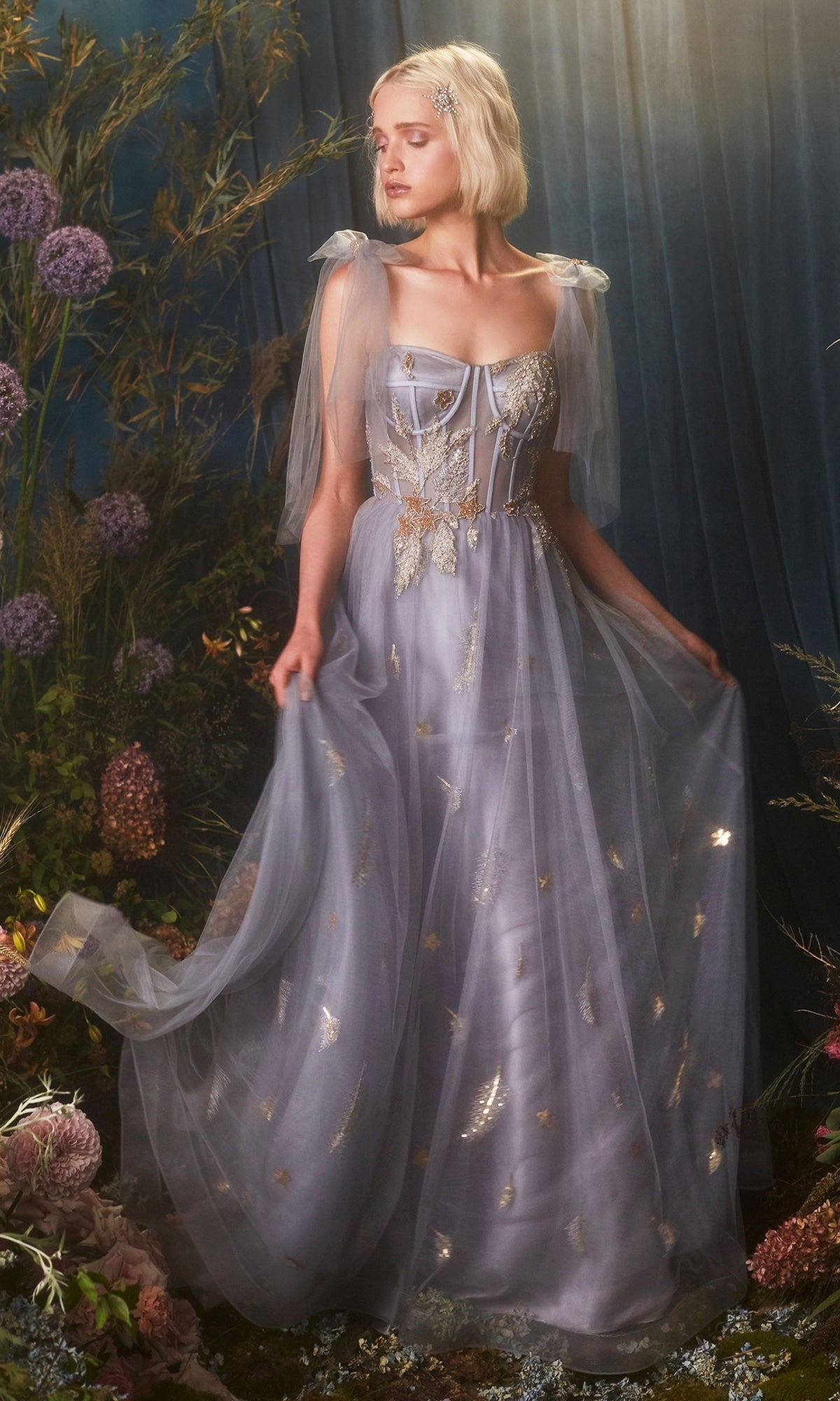 A woman wears the Shoulder-Bow Long Corset Prom Ball Gown A0824, a sheer lavender dress with gold details and tulle sleeves, standing amid lush florals and dark draped curtains.